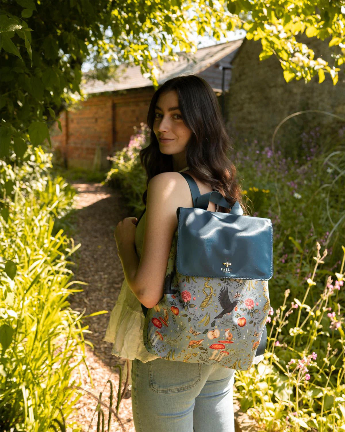 A women with long browA woman sporting the teal Into the Woods backpack, featuring a cottagecore design by Catherine Rowe. The print on backpack has crows, mushrooms, flowers, and other nature motifs.